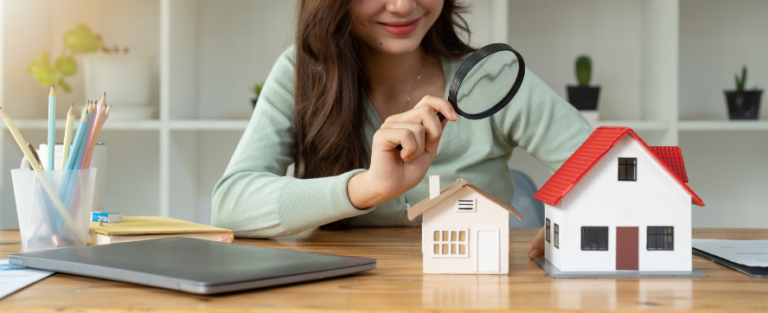 Student sitting at a desk with a model house in front of her and she is inspecting the model house with a magnifying glass. This is to represent her searching for a home. There are also usual items that students use daily such as her laptop and pencils on the desk.