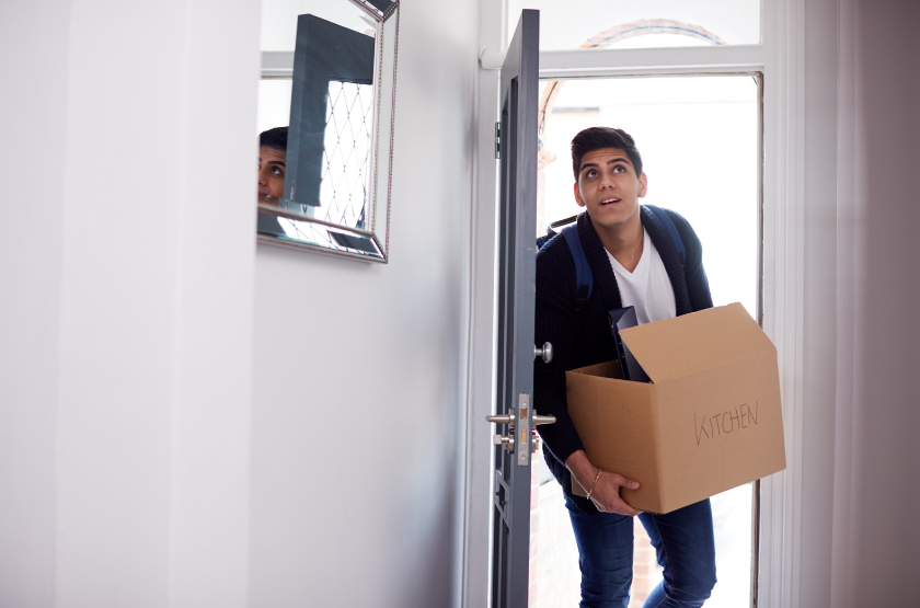 Male Student Walking into a property through a white door and carrying a cardboard box with the label kitchen written across it. He looks curious yet excited as he is walking into the home and is stood in the doorway and keeping the door open to by holding it with his body.