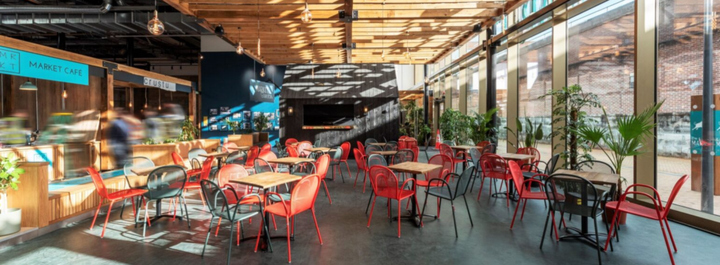 An image of the market at Chester where there is seating and you can see the stand alone stalls where food can be purchased. There are lots of windows letting in natural light, exposed bulbs that are hanging down to replicate the industrial setting that Chester Market is within and plants that make it the garden room