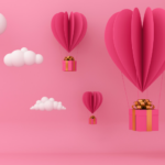 Pink background with darker pink heart shaped paper decorations that are mimicking hot air balloons. White clouds are surrounding them.