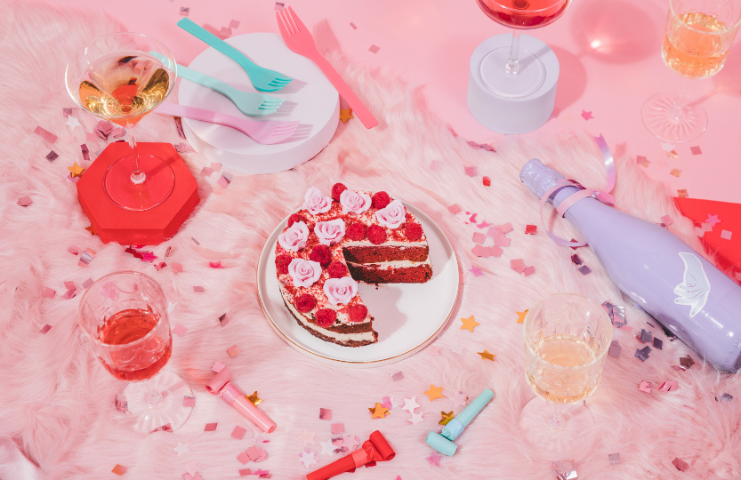 An image that is representing a girls night out celebrating valentines together. There are items on a pink background that represent some things that are associated with a girls night out usually, such as glasses of different drinks, there is also a cake that has had a slice cut out of it that has rose petals on the top as decorations and heart shaped ones to represent it is a nod to valentine's day. There is confetti shaped as starts that is shiny representing that there has been a celebration or party.