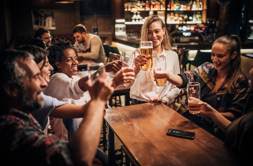 Lots of younger people stood around a table within a bar holding drinks and all smiling at one another. They are holding drinks towards each others drinks as though they are celebrating.