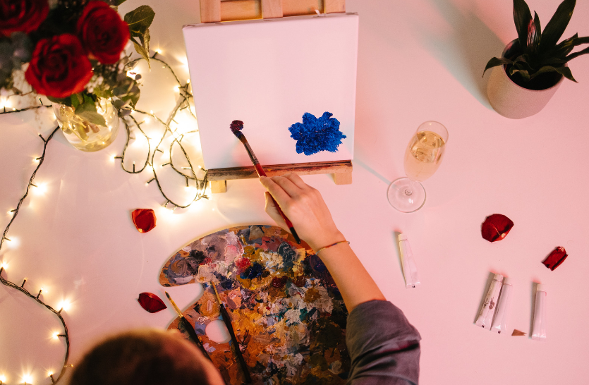 This image is portraying a person taking part in an activity which is them painting a canvas. There is a vase of red roses to the left and some fairy lights. The roses are indicating it is a romantic setting like Valentine's and the fairy lights portray the image of intimacy.
