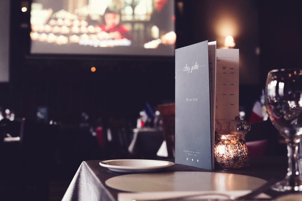 An image of the interior of chez jules, it is in a dark room that is lit with dimly lit lighting making the room feel intimate and cosy. There is a table that is set with a drinks menu on it and there is a screen in the background that is distorted showing where the cinema showing is.